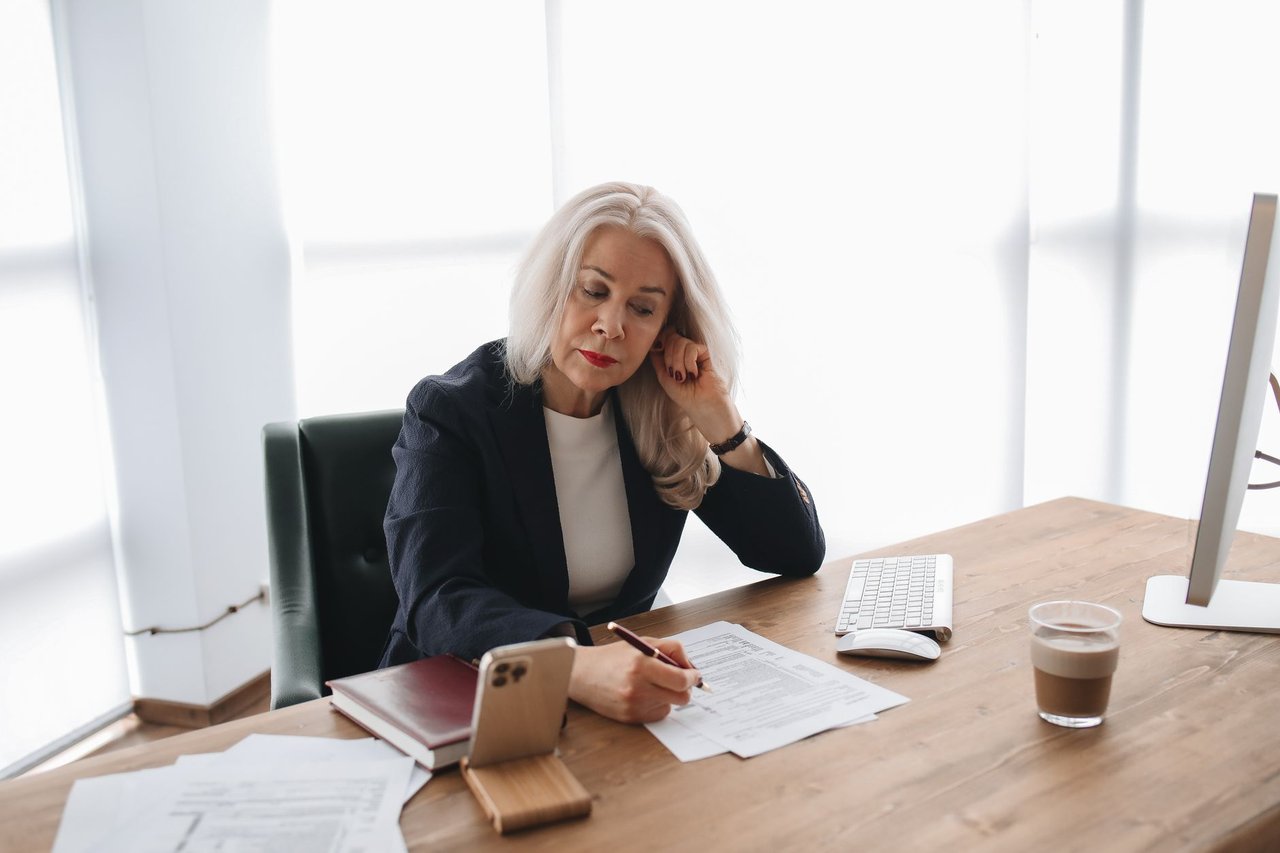 Profissional revisa documentos em uma mesa de escritório ao lado de um computador e um smartphone.