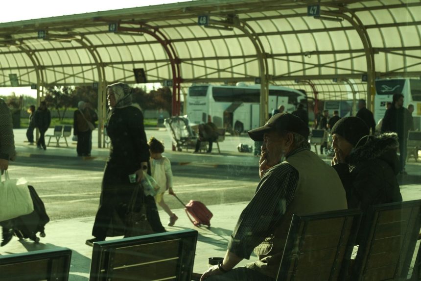 Passageiros circulam e aguardam em um movimentado terminal rodoviário ao ar livre.